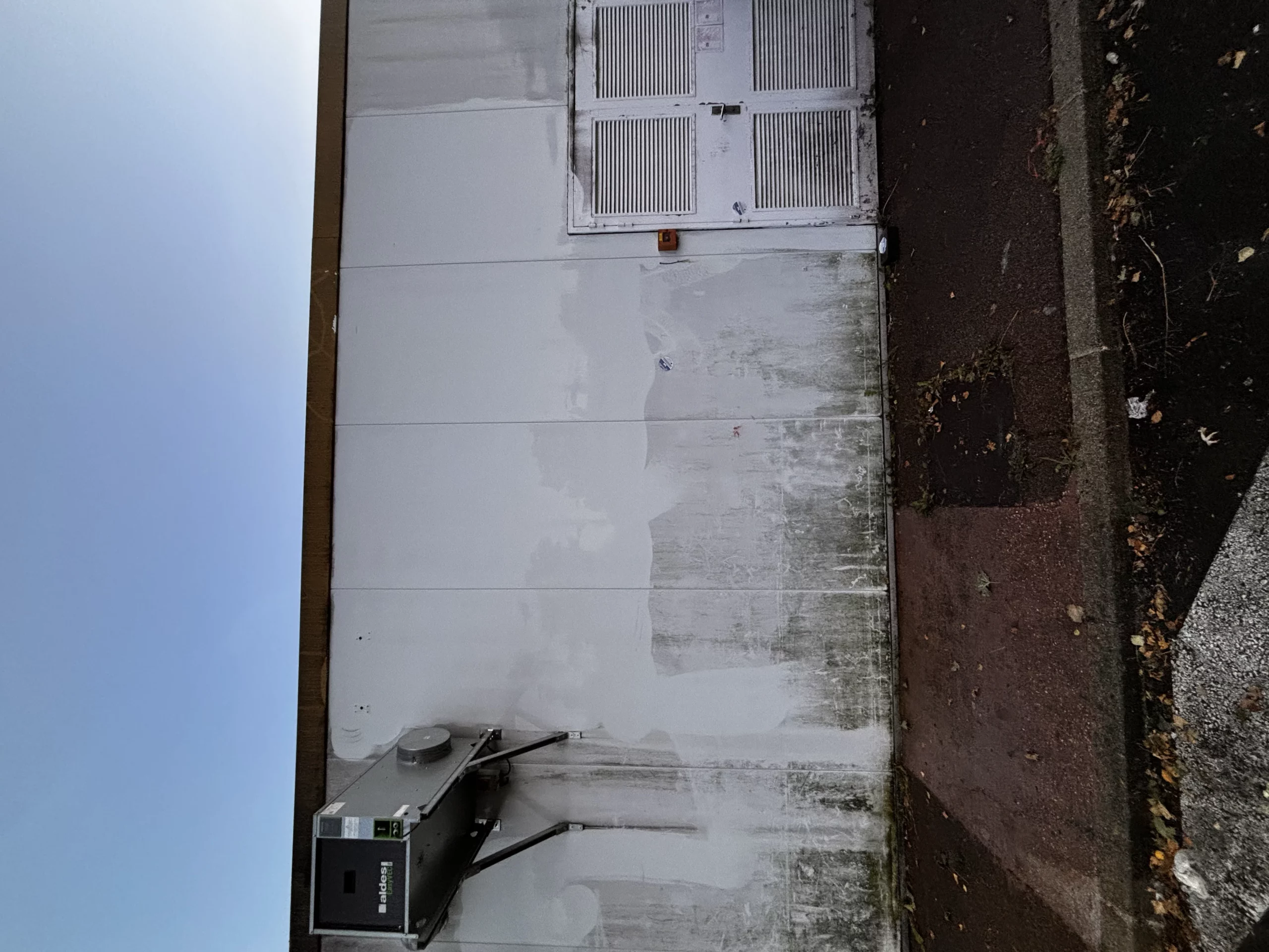 Mur extérieur blanc avec boîtier technique, panneaux de ventilation et passage pavé le long du bâtiment.